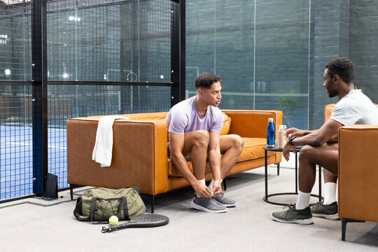 Diverse male friends in athletic wear resting in padel lounge with racket, ball, coffee cup