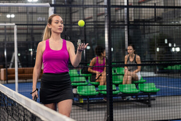 Diverse female teammates tossing tennis ball with padel racket next to net and mesh fence