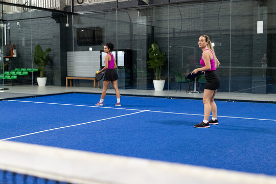 Diverse female teammates in fuchsia outfits playing padel on blue court with rackets, ball
