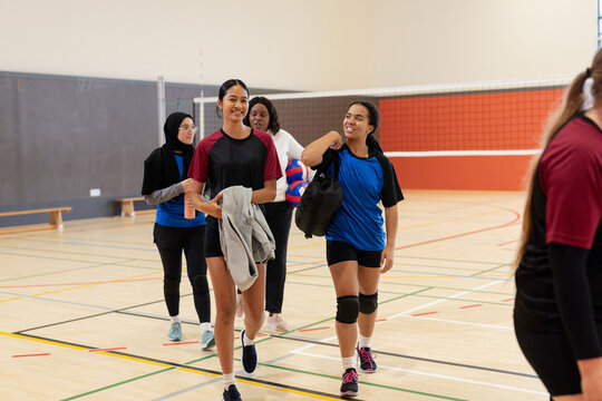 Diverse female teammates walking across court by volleyball net with gym bag and volleyballs