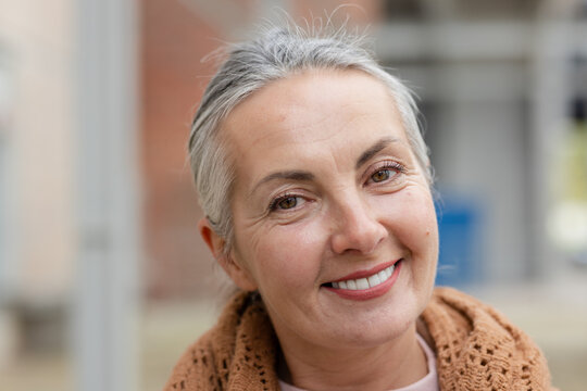 Senior woman smiling, wearing brown knitted shawl over pale pink top in blurred urban courtyard
