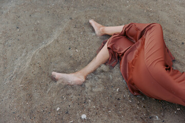 Bare legs stretch on a sandy, pebbled surface, a rust colored dress draped around the body. The scene captures stillness, warmth, and soft summer light, with quiet texture.