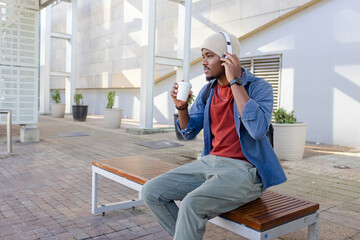 African american male sipping coffee and adjusting headphones sitting on plaza bench, backpack