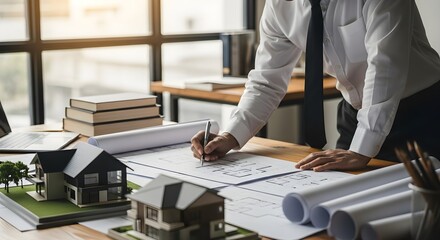 Architect working on blueprints at a desk with house models and rolled plans, wearing a suit and tie, in a bright office setting.
