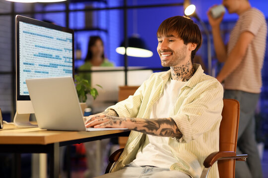 Tattooed male programmer working with laptop in office at night