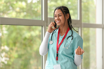 Young African-American female doctor with stethoscope and badge talking by mobile phone near window in clinic