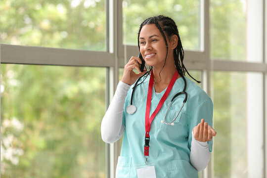 Young African-American female doctor with stethoscope and badge talking by mobile phone near window in clinic