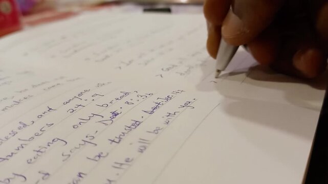 African sitting, writing with white pen on white paper sheet page with black cover diary on table. Having Bible studies, taking notes. Blue ink handwritten, written.