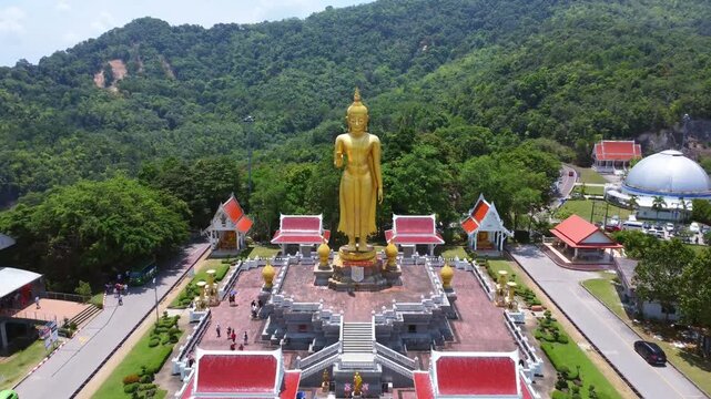Drone video of towering golden Buddha on raised platform, surrounded by vibrant red-roofed temples amid lush forested Mae Hong Son hillside. Clear, bright lighting accentuates vibrant colors.