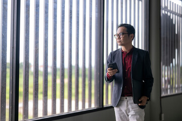 Pensive businessman holding smartphone and laptop by office window