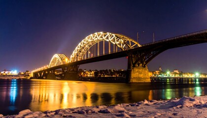 Naklejka premium Illuminated bridge spanning a river at night, its lights reflected in the calm water, snow visible on the banks