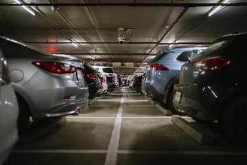 A dimly lit underground parking lot filled with cars parked in rows, featuring security cameras, concrete flooring, and overhead fluorescent lights.