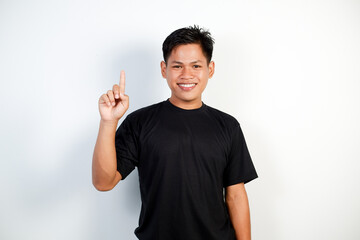 Fototapeta premium A Cheerful Young Asian Man Wearing a Black Shirt Pointing Upward With a Smile on a White Background.