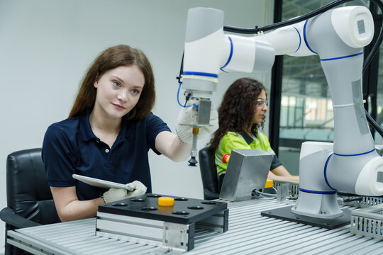 female technician wearing navy shirt and gloves practicing precision control of robotic arm during automation skills training session in smart factory environment using tablet and machinery
