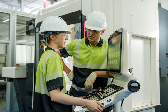 Female and male technicians operating CNC machine interface during smart factory training session wearing safety helmet and glasses collaborating on precision control and programming development