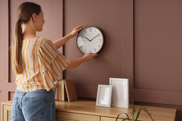 Young woman hanging clock on brown wall in room, back view