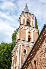 A stunning timeless landmark view of a Georgian Orthodox church bell tower with a pointed spire, elegant arched windows, and detailed brickwork. Surrounded by lush green trees