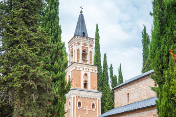 Obraz premium A scenic view of a Georgian Orthodox church featuring its tall brick bell tower with a pointed spire, framed by lush green cypress trees. This historic landmark reflects the timeless beauty of Georgia