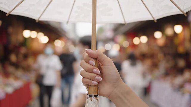 Woman's Hand Holding Parasol in Busy Market - Close-up vertical shot of a woman's hand holding a white parasol with a subtle pattern.