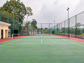 Empty tennis court with green and red surface, surrounded by tall wire fencing under an overcast sky, creating a quiet atmosphere for practice, competition, or leisure play in a peaceful outdoor setti