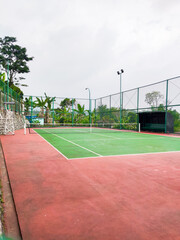 Empty tennis court with green and red surface, surrounded by tall wire fencing under an overcast sky, creating a quiet atmosphere for practice, competition, or leisure play in a peaceful outdoor setti