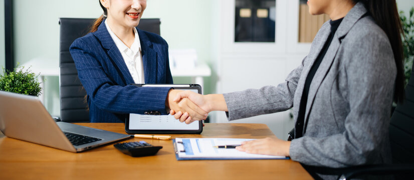 Two female professionals shake hands over financial reports and contract papers in an office, symbolizing successful business agreement, teamwork, and corporate partnership.