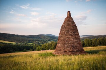 Tall, conical woodpile in a grassy field, hills in the background