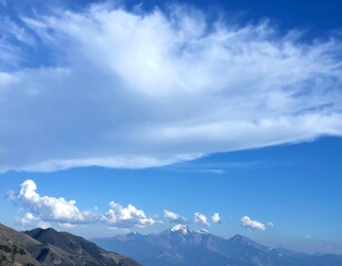A vast, clear blue sky, dotted with soft, fluffy clouds, overlooks a mountain range, showcasing a serene and tranquil landscape.