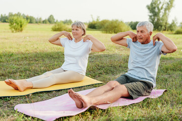 Active elderly couple doing crunches on yoga mats in a field.