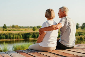 Mature couple embracing and looking at a river, sitting on a wooden pier during sunset.