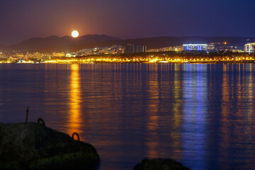 Obraz premium Gelendzhik resort at night by moonlight. Lights reflected in Gelendzhik Bay. Caucasus Mountains in background. Stones in foreground.