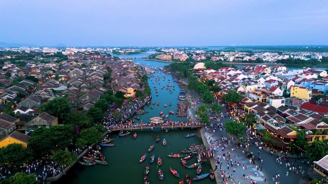 Aerial view of hoi an historic district, colorful boats drifting along thu bon river, pedestrian bridge silhouetted against twilight sky, unesco world heritage landmark in vietnam