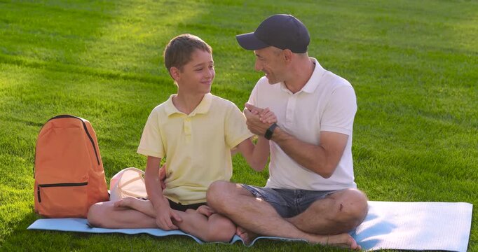 Happy father and son bonding outdoors on sunny park lawn at sunset. Smiling dad and child sit together on picnic blanket in nature, sharing handshake shows trust, love, and support.Warm family moment.