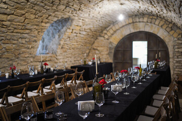Stone cellar dining hall with long tables, wine glasses, candles, and red flowers creating a rustic romantic atmosphere