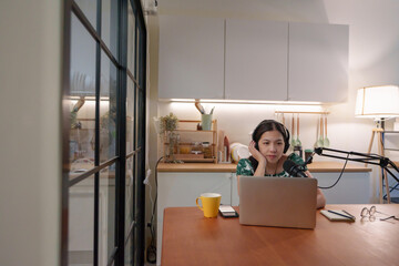 Asian female podcaster records audio at desk with microphone and laptop, working from home on digital content.