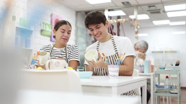 Happy young guy and girl spending quality time together in pottery studio, creating and decorating ceramic cups. Creativity and bonding concept