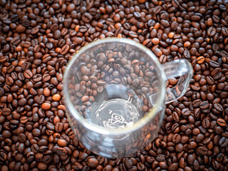 Empty glass coffee cup placed on roasted coffee beans. Transparent mug blends with background of brown beans. Concept of coffee, cafe and caffeine.