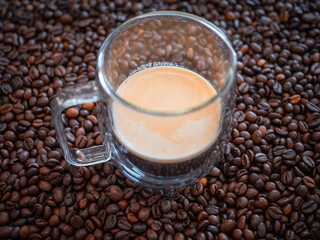 Glass cup filled with espresso coffee placed on roasted coffee beans background. Fresh hot drink with crema foam, concept of caffeine, breakfast and cafe design.