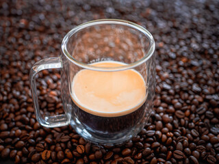 Glass cup filled with espresso coffee placed on roasted coffee beans background. Fresh hot drink with crema foam, concept of caffeine, breakfast and cafe design.