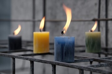 Four burning candles in various colors (yellow, blue, dark-gray, green) on a metal grate against a light gray wall