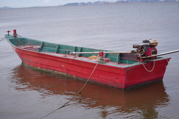 A red long-tail boat floating on calm seawater under a partly cloudy sky, with distant islands on the horizon, capturing the peaceful simplicity of coastal fisherman life
