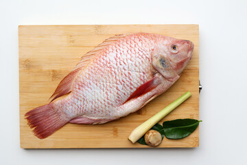Fresh Raw Red Tilapia fish and Galangal, lemongrass, kaffir lime leaves on a wooden board on a white background.