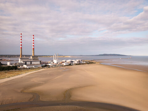 Aerial view of the Poolbeg Generating Station's red and white chimneys standing tall against the coastal landscape, with the sea gently lapping the sandy shore, Dublin, County Dublin, Ireland.