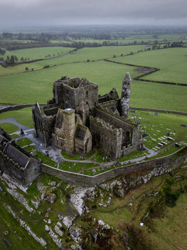 Aerial view of the Rock of Cashel's formidable stone ruins piercing the vibrant green landscape under a moody sky, Cashel, County Tipperary, Ireland.