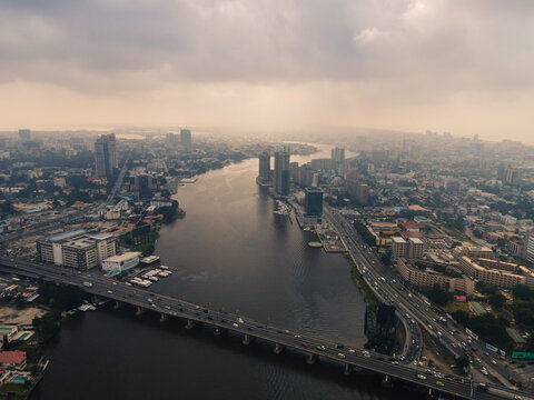Aerial view of the bustling Ozumba Mbadiwe Avenue bridge connecting to high-rise buildings across the still, dark water under a pale sky, Lagos, Lagos, Nigeria.