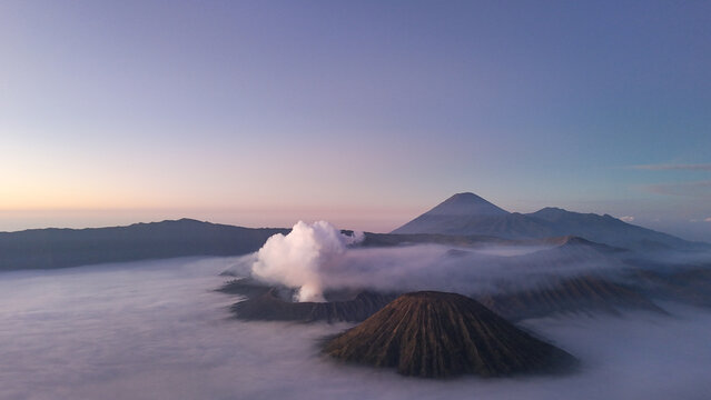 Aerial view of Mount Bromo volcano at sunrise surrounded by clouds and mist in East Java, Indonesia. Scenic volcanic landscape with smoke, mountains, and dramatic sky.