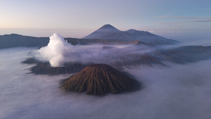 Aerial view of Mount Bromo volcano at sunrise surrounded by clouds and mist in East Java, Indonesia. Scenic volcanic landscape with smoke, mountains, and dramatic sky.