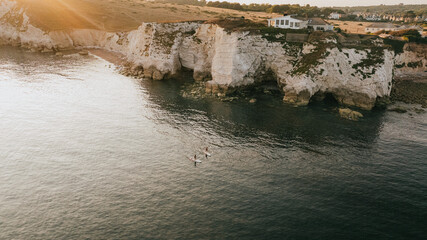 A couple of people are paddling in the ocean near a house. The water is calm and the sky is orange