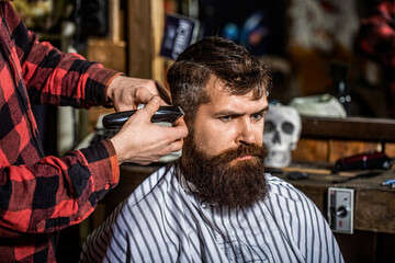 Obraz premium Barber works with hair clipper. Hipster client getting haircut. Hands of barber with hair clipper. Bearded man in barbershop. Man visiting hairstylist in barbershop