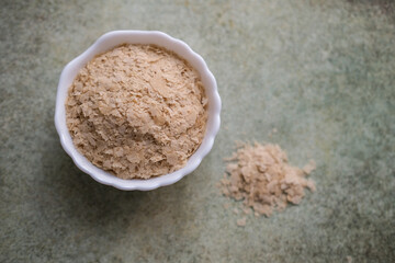 Overhead shot of nutritional yeast flakes in a small white bowl with small pile of flakes next to it on speckled background with copy space